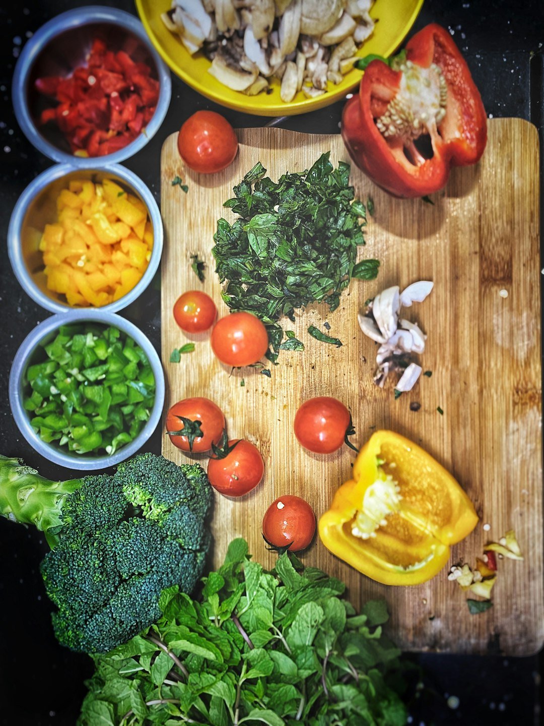 Fresh peppers, tomatoes, broccoli, herbs, garlic, and mushrooms arranged around a wooden cutting board